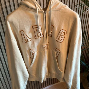 Arigato hoodie - Snygg beige hoodie med dragkedja och stora broderade bokstäver framtill. Tröjan har huva med snörning och en stor magficka. Perfekt för en avslappnad och trendig look.