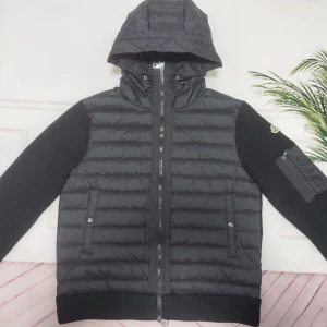 Svart Moncler pufferjacka med huva - Svart pufferjacka från Moncler med stickade ärmar och vadderad kropp. Jackan har huva med snörning, dragkedja framtill och praktiska fickor. Ärmen har en extra ficka med dragkedja och Moncler-logga. Perfekt för kyliga dagar med sin kombination av stil och funktion.