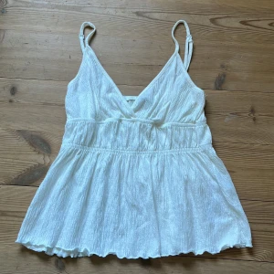 Offwhite babydoll top Gina Tricot - Supersöt offwhite babydoll topfrån Gina Tricot med smala justerbara axelband och omlott-v-ringning. Toppen har markerad midja med rynk och lätt utställd nederdel. Perfekt för varma dagar och enkel att styla med jeans eller kjol. Prislappen finns kvar, helt ny