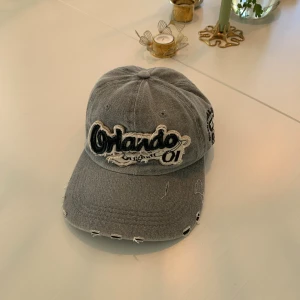 Grå keps med Orlando-brodyr - Snygg grå keps med broderad Orlando-text framtill och detaljer på sidan. Kepsen har slitna kanter och coola broderier som ger en vintagekänsla. Justerbar baktill för perfekt passform. 