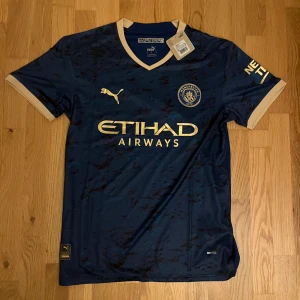 Manchester City bortatröja Puma blå - Manchester City bortatröja från Puma i mörkblått med guldiga detaljer och klubbmärke på bröstet. Tröjan har korta ärmar, V-ringning och är tillverkad i lätt funktionsmaterial. Snyggt mönster över hela framsidan och kontrastfärgade muddar.