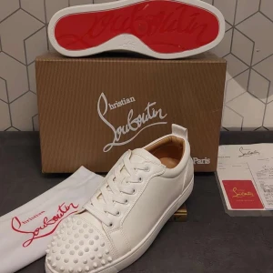 Christian Louboutin vita sneakers med nitar - Säljer ett par vita sneakers från Christian Louboutin med ikonisk röd sula och coola nitar framtill. Skorna är i skinn och har snörning. Perfekta för dig som vill sticka ut med en lyxig och edgy stil.