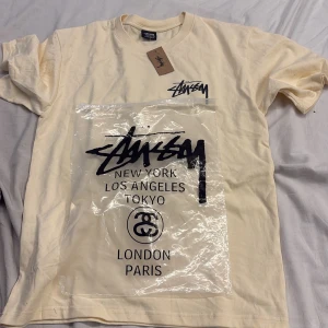 Stüssy t-shirt - Snygg beige t-shirt från Stüssy med svart logga och tryck på bröstet samt stor grafisk print med blå tärningar på ryggen. Klassisk rund hals och korta ärmar. Perfekt för dig som gillar streetwear och vill sticka ut med coola detaljer. Jag kan inte garantera äktheten på denna vara.