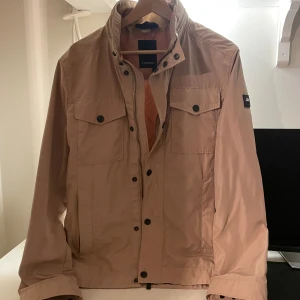 Beige vindjacka från J.Lindeberg M - Stilren beige vindjacka från J.Lindeberg i storlek M. Jackan har två bröstfickor med knappar, hög krage och diskret logotyp på ärmen. Tillverkad i lätt polyester, perfekt för vår och höst. Snygga detaljer med svarta knappar och dragkedja framtill.