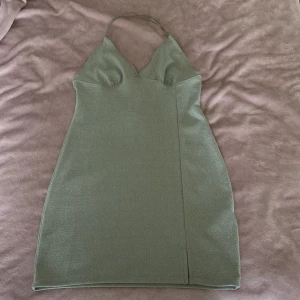 Grön glittrig kort klänning H&M Divided - Snygg grön glittrig kort klänning från H&M Divided i storlek M. Klänningen har smala axelband som korsas i ryggen, v-ringad hals och en liten slits framtill. Materialet är stretchigt och skimrande, perfekt för att sticka ut.