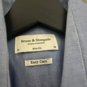 Blå skjorta från Bruun & Stengade - Slim fit skjorta i ljusblått från Bruun & Stengade. Klassisk krage, knäppning framtill och långa ärmar. Skjortan är tillverkad i ett mjukt bomullstyg och har Easy Care-finish som gör den enkel att sköta. Perfekt för en clean och stilren look. Aldrig använd.