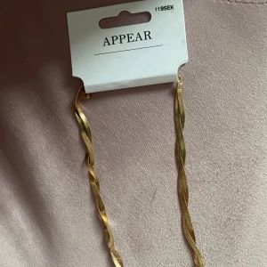 Guldigt tvinnat halsband från Appear - Säljer ett snyggt guldigt halsband från Appear med tvinnad design. Kedjan har en elegant vågig form som ger en modern och trendig look. Perfekt accessoar för att lyfta vilken outfit som helst.