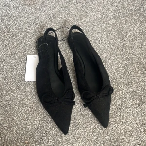 Svarta slingbacks med rosett H&M - Svarta slingback från H&M med spetsig tå och dekorativ rosett framtill. Skorna har öppen häl och en elegant, tunn rem runt ankeln. Tillverkade i syntetmaterial med en platt sula, perfekta för en stilren look. Nyköpta för 299
