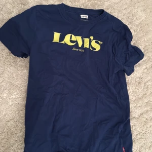 Blå Levi's t-shirt med gul logga - Snygg marinblå t-shirt från Levi's med stor gul logga på bröstet. Klassisk passform och rund hals. Tillverkad i 100% bomull för en skön känsla. Perfekt för dig som gillar stilrena och ikoniska plagg.