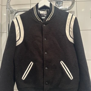 Svart bomberjacka från Saint Laurent - Säljer en svart och vit bomberjacka från Saint Laurent Paris. Jackan har klassisk collegekrage med ränder, vita detaljer på axlar och fickor samt knäppning framtill. Materialet är ullblandning med fodrad insida. Perfekt för dig som vill ha en stilren och lyxig look.
