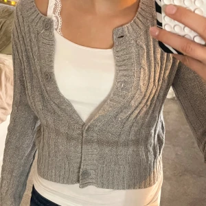 Grå kofta med kabelstickning - Fin Grå kofta med kabelstickat mönster och stora knappar framtill. Koftan har lång ärm och ribbade muddar vid ärmslut och nederkant. Koftan är lite kort för mig då den ser lite ”croppad” ut men skulle säga att den är normallång.