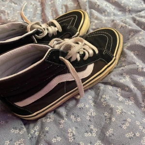 Svarta Vans Sk8-Hi sneakers - Klassiska Vans Sk8-Hi sneakers i svart med vit sula och vit sidostrip. Skorna har snörning, hög modell och är tillverkade i mocka och canvas med detaljer i vitt skinn. Perfekta för en streetwear-look och passar till det mesta.