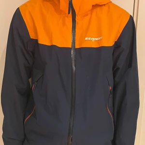 Blå, orange regnjacka Everest - Snygg tvåfärgad regnjacka från Everest, Stadium, i mörkblått och orange. Jackan har huva, hel dragkedja framtill och praktiska fickor med dragkedja. Logga på bröstet och meshfoder på insidan.