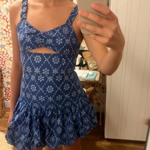 Blå blommig kortklänning med cut-out - Supersöt blå kortklänning med vita blommor och rutmönster. Använd endast en gång!. Klänningen har breda axelband, cut-out detalj framtill och volangig kjol. Perfekt för varma dagar och ger en riktigt somrig vibe! 