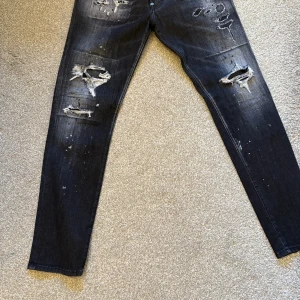 Svarta Dsquared2 jeans med slitningar - Riktigt snygga svarta jeans från Dsquared2 med slitna detaljer och färgstänk. Modellen har smal passform och stort Dsquared2-tryck på bakfickorna. Jeansen är tillverkade i Italien och har en cool, edgy vibe som sticker ut.