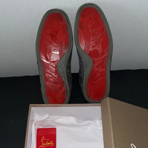 Christian Louboutin sneakers med röd sula - Exklusiva sneakers från Christian Louboutin med ikonisk röd sula och grå mocka på ovandelen. Skorna har rund tå, snörning och lyxig läderinsida. Kommer med originalkartong och dustbag. Perfekta för dig som vill sticka ut med stil. Den kommer att få en skönare tvätt innan varan skickas! 