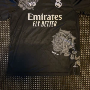 Real Madrid Y-3 120-års jubileumströja - Snygg svart Real Madrid fotbollströja från Y-3 kollektionen, designad för klubbens 120-årsjubileum. Tröjan har korta ärmar, stora gråa rosor som tryck och klassiska loggor framtill. Tillverkad i lätt funktionsmaterial (AEROREADY) för maximal komfort.