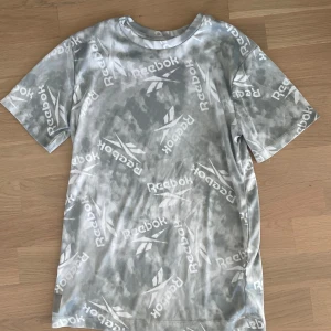 Grå mönstrad Reebok t-shirt - Snygg grå t-shirt från Reebok med coolt mönster och logga tryckt över hela plagget. T-shirten har rund hals och korta ärmar, perfekt för dig som gillar sportig stil. Materialet är mjukt polyester som känns skönt mot huden.