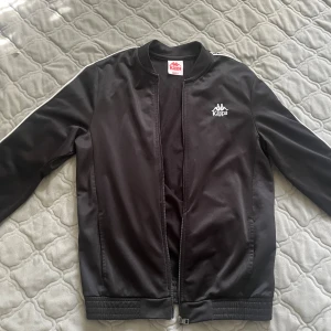 Svart Kappa bomberjacka med logoband - Svart bomberjacka från Kappa med klassiska vita logoband längs ärmarna och broderad logga på bröstet. Jackan har dragkedja framtill, ribbade muddar och en sportig vibe. Tillverkad i polyester och har lång ärm. Perfekt för dig som gillar streetwear.kommer inte ta return.