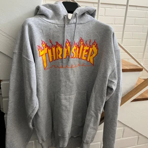 Grå Thrasher hoodie med flames - Säljer en grå hoodie från Thrasher med ikoniskt flames-tryck i gult och rött på bröstet. Tröjan har huva med snörning, magficka och är i mjuk bomullsblandning. Perfekt för dig som gillar streetwear och skatekänsla.
