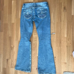 Ljusblå bootcut jeans med broderade fickor - Säljer ett par ljusblå bootcut jeans med låg midja och slitningar på benen. Baksidan har snygga broderade fickor med knappar. Jeansen är i klassisk denim och har en riktigt skön passform som ger en avslappnad vibe.