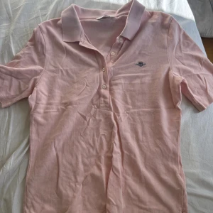 Ljusrosa pikétröja från Gant - Ljusrosa pikétröja från Gant med klassisk krage och knäppning framtill. T-shirten har korta ärmar och liten broderad logga på bröstet. Tillverkad i mjuk bomull som känns skön mot huden. Perfekt för en avslappnad och stilren look.