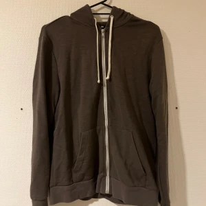 Brun hoodie med dragkedja från H&M - Mysig brun hoodie från H&M med vit dragkedja och snörning i huvan. Tröjan har två fickor framtill och är långärmad med ribbade muddar. Perfekt för chill dagar eller när du vill ha något bekvämt och enkelt.