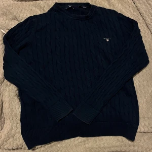 Marinblå kabelstickad tröja GANT - Snygg marinblå kabelstickad tröja från GANT med rund hals och diskret logga på bröstet. Tröjan har långa ärmar, ribbade muddar och är gjord i mjuk bomull. Perfekt för dig som gillar klassisk stil med en modern touch (den är oanvänd). Kan vara lite stor i storleken så skulle precis kunna passa en person som är 170 men för bäst passform mellan 158-164 mer eller mindre.