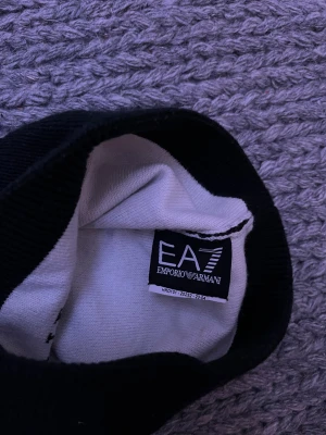  EA7 Emporio Armani mössa - Svart mössa från EA7 Emporio Armani med stor vit logga framtill. Klassisk ribbad kant och mjukt material som sitter skönt på huvudet. Perfekt accessoar för att hålla värmen och ge din outfit en lyxig touch.