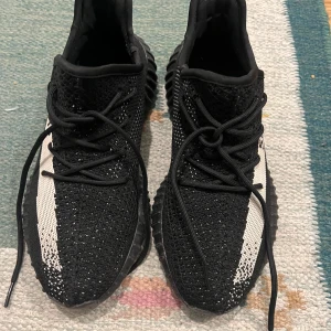 Adidas Yeezy Boost 350 V2 svart/vit - Säljer ett par Adidas Yeezy Boost 350 V2 i svart och vit med SPLY-350 text på sidan. Skorna har stickad ovandel i syntetmaterial, snörning och en platt sula med ribbad struktur. Perfekt för dig som gillar streetwear och vill ha något unikt på fötterna.