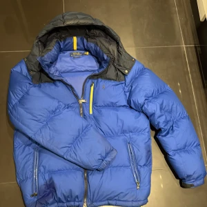 Blå dunjacka Polo Ralph Lauren - Blå dunjacka från Polo Ralph Lauren med mörkgrå detaljer på axlar och huva. Jackan har flera dragkedjeförsedda fickor, gul detalj vid kragen och är fodrad med dun och polyester. Perfekt för kalla dagar och har en sportig vibe. OBS jackan har även en liten lagning på höger arm kan skicka bild bara att skriva! Priset är ej hugget i sten.