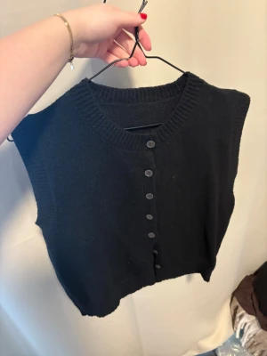 Svart stickad väst med knappar - Svart stickad väst med rund hals och ribbade kanter. Västen har en rad svarta knappar framtill och är ärmlös, vilket gör den enkel att styla över en t-shirt eller skjorta. Perfekt för lager-på-lager och en clean look.