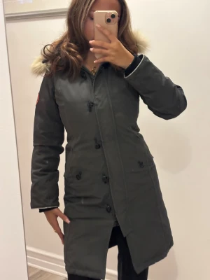 Canada Goose mörkgrå parkas med päls - Säljer en snygg mörkgrå parkas från Canada Goose med fluffig päls på huvan och klassisk logga på ärmen. Jackan har långa ärmar, knappar framtill och två fickor. Perfekt för kalla dagar och riktigt stilren look.