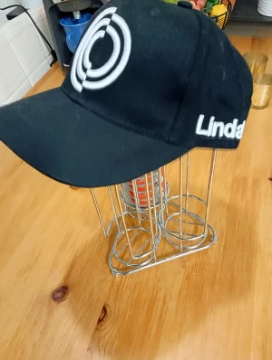 Svart keps från Lindab med broderi - Svart keps med böjd skärm och vit broderad logga framtill. Snyggt Lindab-tryck på sidan. Klassisk modell som passar till streetwear och chill vardagsstil. Tillverkad i bomull för skön känsla och bra passform.