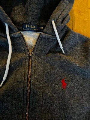 Mörkgrå hoodie från Polo Ralph Lauren - Snygg mörkgrå hoodie från Polo Ralph Lauren med vit snörning i huvan och röd broderad logga på bröstet. Tröjan har dragkedja framtill, två fickor och är tillverkad i mjuk bomullsmix. Perfekt för chill dagar och streetstyle.