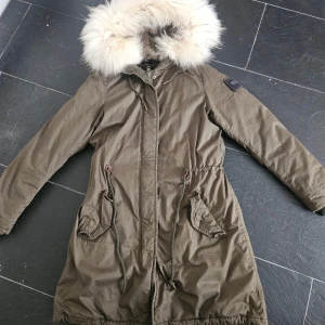 Olivgrön parkajacka med päls från The Smirk - Parkajacka i olivgrönt från The Smirk, New York. Jackan har en stor fluffig huva med ljus päls, snörning i midjan och två praktiska fickor framtill. Yttertyget är i bomull och fodret i polyester, perfekt för kalla dagar.