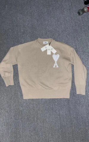 Beige stickad tröja från Ami - Snygg beige stickad tröja från Ami Alexandre Mattiussi med vit hjärtapplikation och A på bröstet. Tröjan har rund hals, ribbade muddar och lång ärm. Perfekt för dig som gillar stilrena och trendiga plagg med cool detalj framtill.