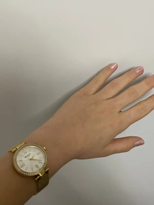 Guldig klocka från Curren med stenar - Ska säljas på grund av storleken som inte passar alls, priset går att diskuteras.                             Snygg klocka från Curren i guld med vit urtavla och romerska siffror. Runt boetten sitter glittrande stenar och armbandet är metall. Klassisk och stilren design som sticker ut med sina detaljer.