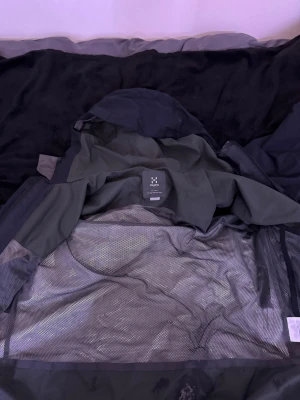 Svart Haglöfs vindjacka med huva - Svart vindjacka från Haglöfs med huva och flera dragkedjeförsedda fickor. Jackan har meshfoder på insidan och är tillverkad i slitstark syntet. Perfekt för utomhusaktiviteter och har en clean, sportig look med diskret logga på bröstet. Lite skrynklig för den har legat i min garderob använd stryk gärn och den är som ny