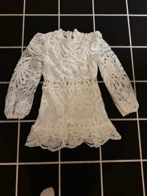 Vit spetsklännibg med volangdetaljer - Superfin vit klänning i spets med långärmad design och volangkant nedtill. Den har ett detaljerat broderat mönster och dragkedja i ryggen. Hög hals och genombrutna partier ger en lyxig och romantisk vibe. Perfekt för dig som gillar statement-plagg!