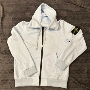 Ljusblå hoodie från Stone Island - Snygg ljusblå hoodie Stone Island med klassisk patch på ärmen. Tröjan har huva med snörning, svart dragkedja framtill och ribbade muddar. Tillverkad i mjuk bomull som är skön att bära. Perfekt för dig som gillar streetwear och vill ha något stilrent.