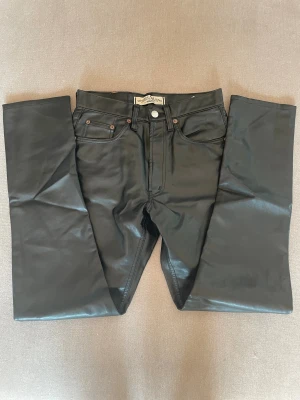 Svarta skinnbyxor från MGB Jeanswear - Svarta byxor i skinnimitation från MGB Jeanswear med klassisk femficksdesign och raka ben. Snyggt läderlook med traditionell jeanspatch bak i midjan. Perfekt för dig som vill sticka ut med en cool och edgy stil. Mått midj tvärs över 33,5 cm, innerben 75 cm