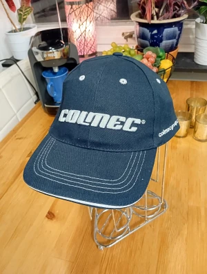 Mörkblå Colmec keps med brodyr - Snygg mörkblå keps från Colmec med vit broderad logga framtill och på sidan. Klassisk form med böjd skärm och vita sömmar som kontrast. Perfekt för dig som vill ha en clean och sportig look.