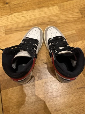 Nike Air Jordan 1 High -Light Smoke Grey 42.5  - Nike Air Jordan 1 High med vit läderbas, grå mockadetaljer och röd skinnpanel vid ankeln. Svarta swoosh-loggor och snörning, samt perforerad tå för extra ventilation. Klassisk high-top siluett med rund tå och platt sula. Perfekt för dig som gillar streetwear och sneakers. Storlek 42.5. Finns tecken på användning som ni ser på bilderna, alltid använt crease skydd. 