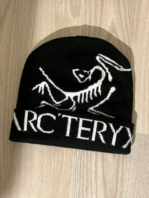 Svart mössa från Arc'teryx - Svart mössa från Arc'teryx med vit logotyp och text framtill. Klassisk form med uppvikt kant och stickad i mjukt material. Perfekt för kalla dagar och snygg att bära till streetwear eller outdoor-stil.