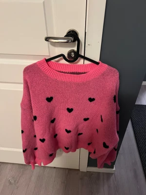 Rosa stickad tröja med hjärtan - Säljer en supergullig rosa stickad tröja med svarta hjärtan över hela. Tröjan har en rund halsringning och långa ärmar med ribbade muddar. Perfekt för dig som gillar färg och söta detaljer. Passar dig som vill sticka ut lite extra!