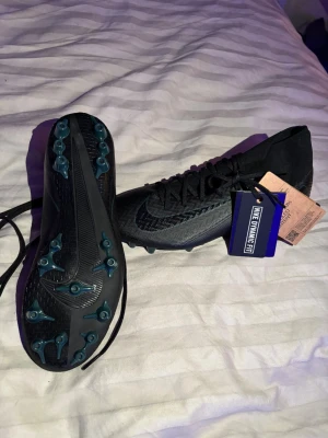 Svarta Nike fotbollsskor med snörning - Säljer ett par svarta Nike fotbollsskor med Dynamic Fit och snörning. Skorna har en högre strumpliknande krage och mönstrad ovandel i syntetmaterial. Sulan har blå dobbar för extra grepp på planen och en stor Nike-logga undertill.