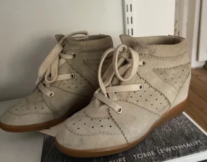 Isabel marant skor - Snygga beige sneakers i mocka med kilklack och platt gummisula. Skorna har snörning framtill, dekorativa perforeringar och vågiga sömmar som ger en cool vintagekänsla. Perfekt för dig som vill ha lite extra höjd men ändå bekvämt. Storlek 39 men passar mig som har storlek 38