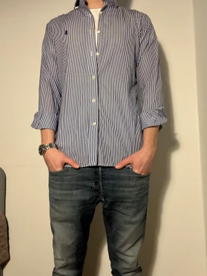 Blåvit randig skjorta från Polo Ralph Lauren - Snygg skjorta från Polo Ralph Lauren med blå och vita vertikala ränder, klassisk krage och knappar framtill. Skjortan har lång ärm och ikoniska Polo-loggan broderad på bröstet. Tillverkad i mjuk bomull för skön känsla och stilren look. Size S 