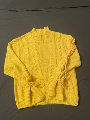 Gul stickad tröja med snörning - Säljer en gul stickad tröja med grova flätstickade detaljer framtill och längs ärmarna. Tröjan har hög hals och cool snörning på båda ärmarna som ger en unik look. Perfekt för dig som gillar färg och vill sticka ut lite extra.